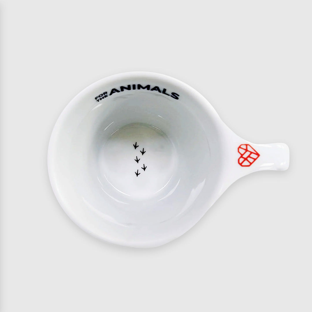View from above the mug showing black chicken footprints in the bottom of the mug. The Mended Heart icon is on the "thumbprint" area above the mug's handle. It is a red heart shape with angular lines within it. For The Animals is written boldly in black on the inner rim of the mug.