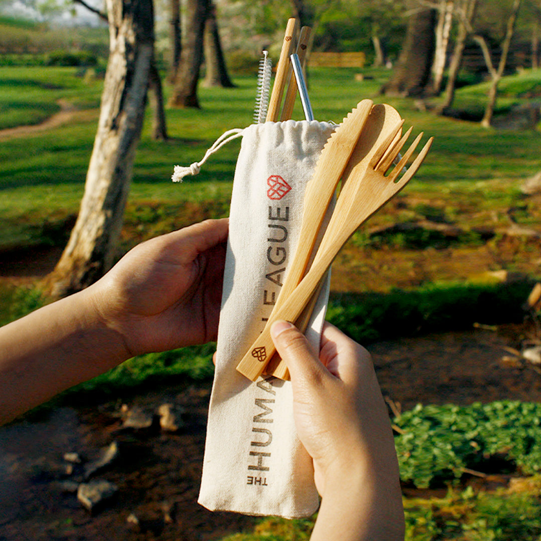 A person holding an eco-friendly bamboo utensil set in an outdoor setting. The utensils, including a fork, knife, spoon, chopsticks, metal straw, and cleaning brush, are stored in a beige drawstring pouch with "The Humane League" printed on it. The background shows a serene, natural landscape with trees and grass.