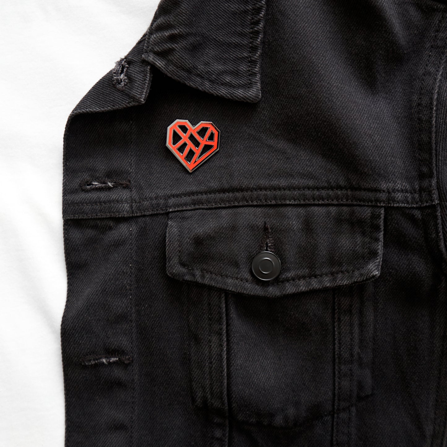 A close-up of a black denim jacket lapel featuring a small geometric heart-shaped pin. The pin has a red enamel fill with silver outlines forming an abstract heart design. Below the pin is a visible chest pocket with a black button closure.