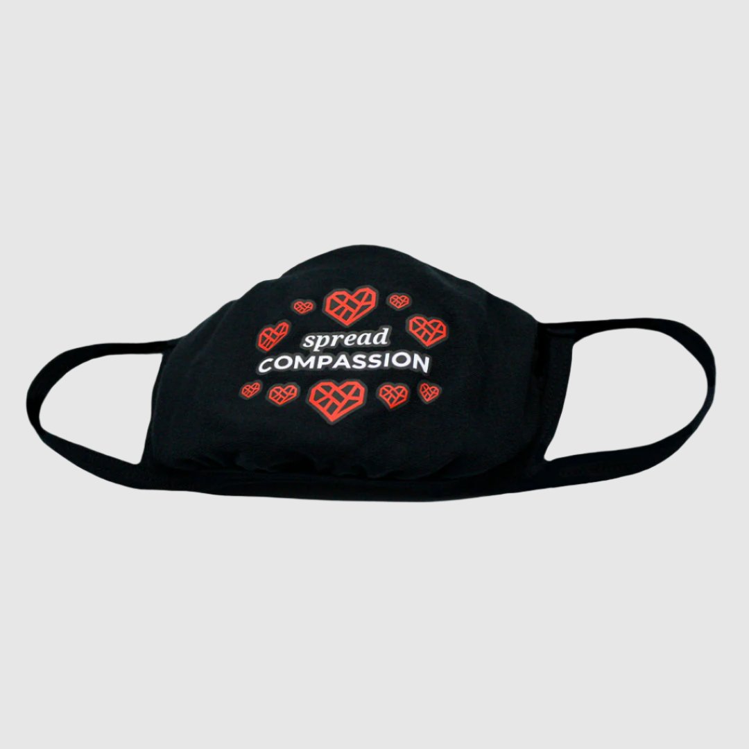 A black fabric face mask with the text "spread COMPASSION" printed in white across the front. Surrounding the text are ten of The Humane League's Mended Heart icons. The mask has ear loops on either side for wearing.