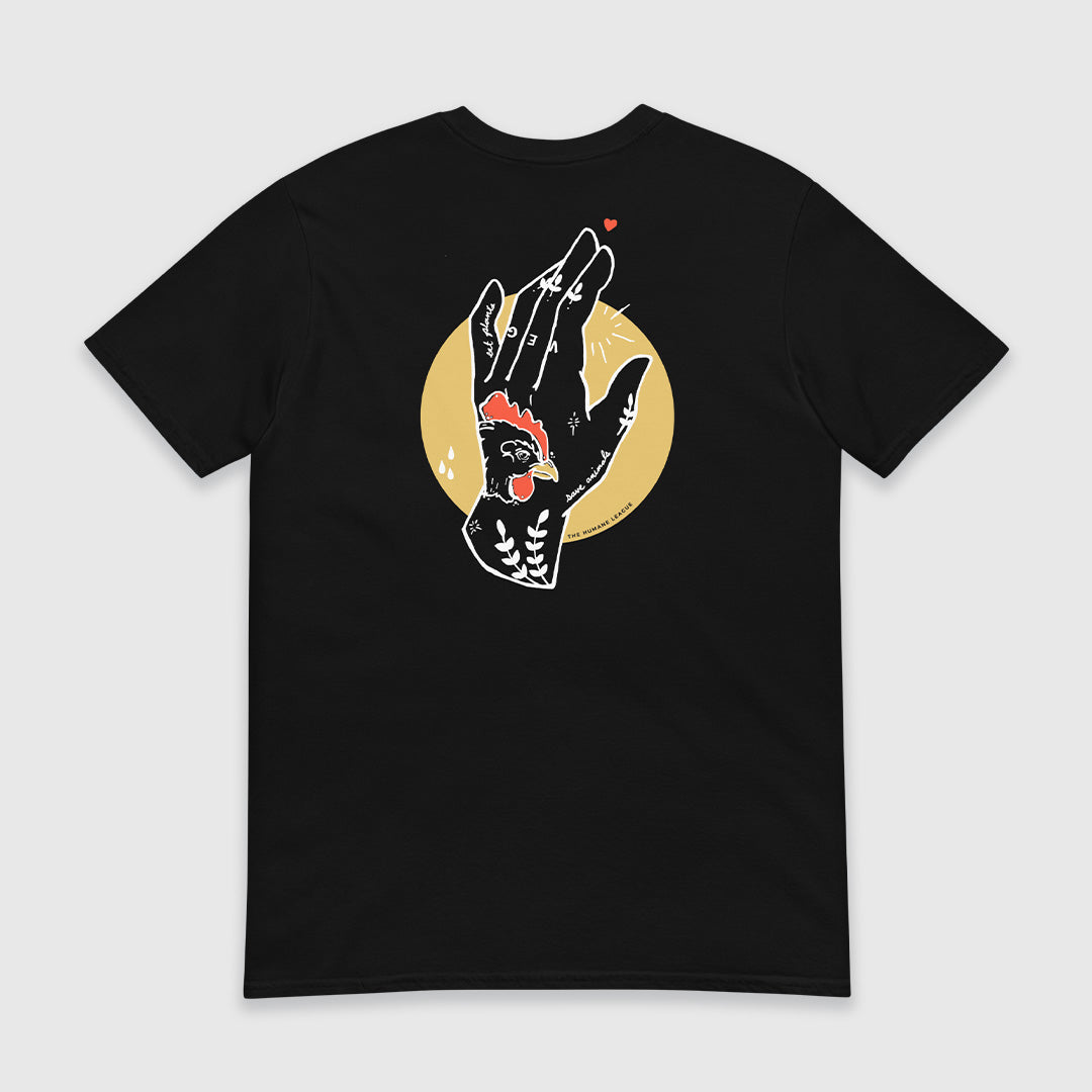 The back of a black short sleeve t-shirt with a design that features a large illustration of an outstretched hand with various tattoos and symbols, including a prominent chicken in red and black on the back center of the hand. White accents and minimalist detailing include some leaves and the words "eat plants" and "save animals". The letters V, E, and G appear written in white on the middle three fingers. The hand is framed within a yellow circle, and a small red heart hovers near the finger tips. 
