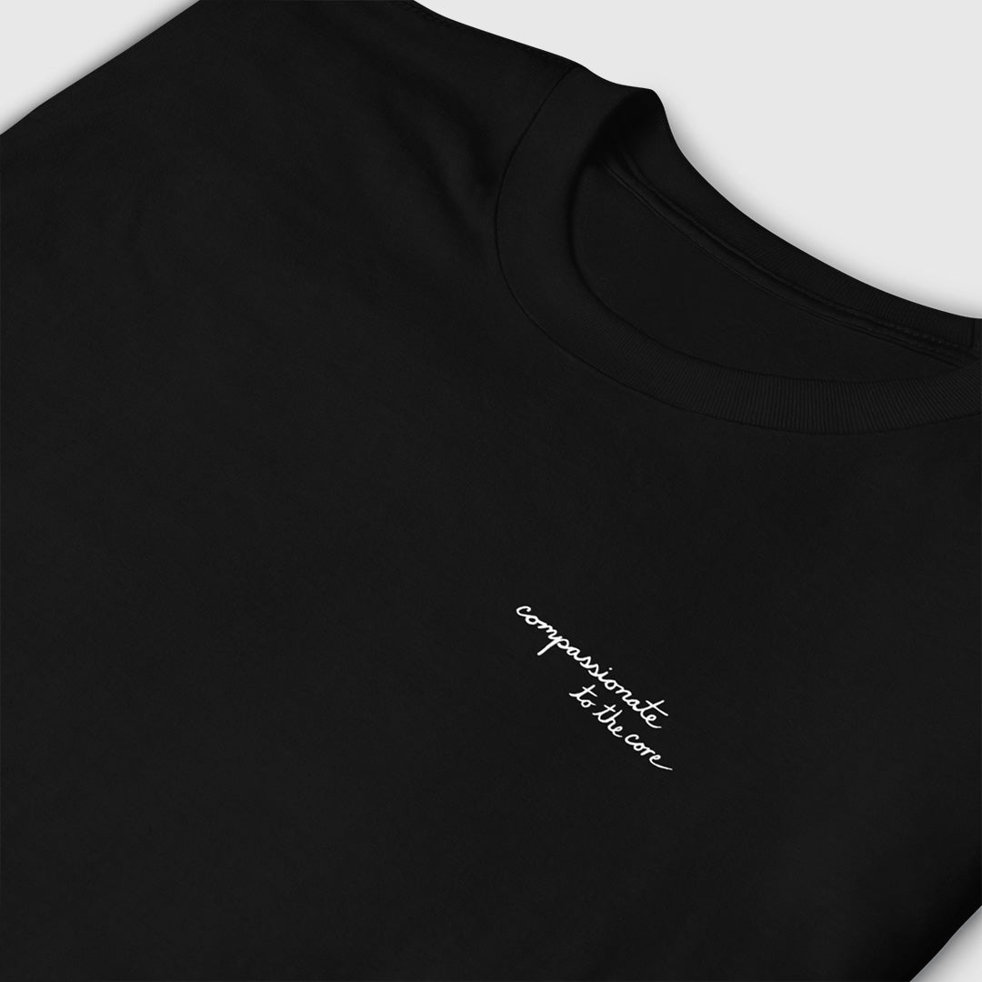 Black t-shirt with close up of white cursive script on right chest area that says, "compassionate to the core".