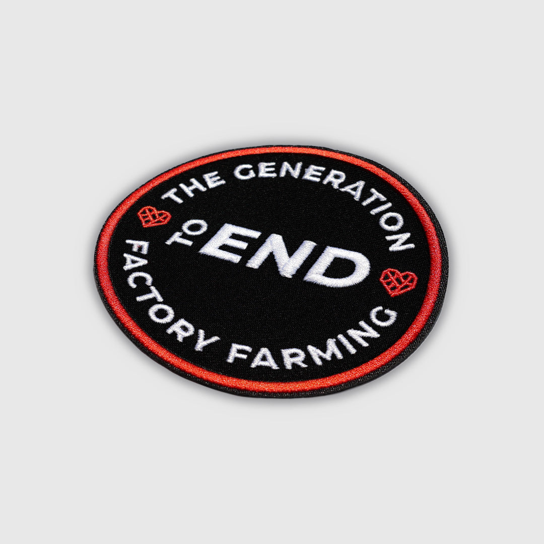 An oval embroidered patch with a black background, red border, and white text reading 'THE GENERATION TO END FACTORY FARMING', with small red heart-like graphics on the sides, shown on a light gray background.