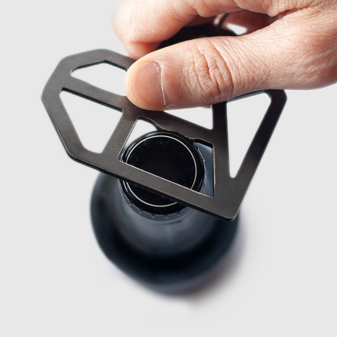 A close-up view of a hand using a geometric heart-shaped metal bottle opener on a dark glass bottle. The bottle opener, made of dark metal with angular cut-outs, is positioned over the bottle cap. A finger is pressing down on the opener to leverage it against the cap. The bottle is partially visible, out of focus in the foreground.