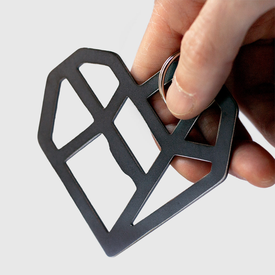 Close-up of a hand holding a black geometric heart-shaped keychain. The heart is made of angular, interconnected lines forming an abstract lattice design. The keychain is attached to a metal ring, which the person's thumb and index finger are gripping. The heart is flat and made of metal. The background is white, and the focus is on the keychain and the fingers holding it.