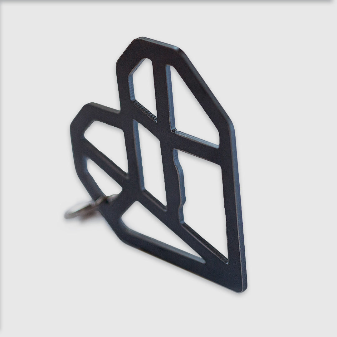 Close-up view of a black geometric heart-shaped keychain or pendant. The heart is made of angular, interconnected lines forming an abstract lattice design. Part of the heart design is a bottle opener. It's shown at an angle, revealing its depth and the clean edges of the cut-out shapes. A small part of a metal key ring is visible attached to the bottom left corner. The object is photographed against a white background, with a shallow depth of field focusing on the front edge of the heart.