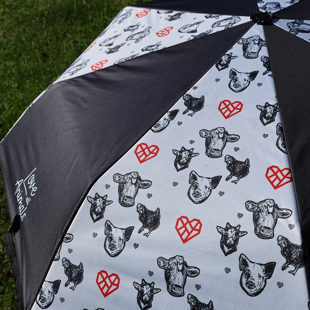 A close-up of a folding umbrella with a black and white background, featuring a playful pattern of cow and pig faces, and chickens, and red hearts.