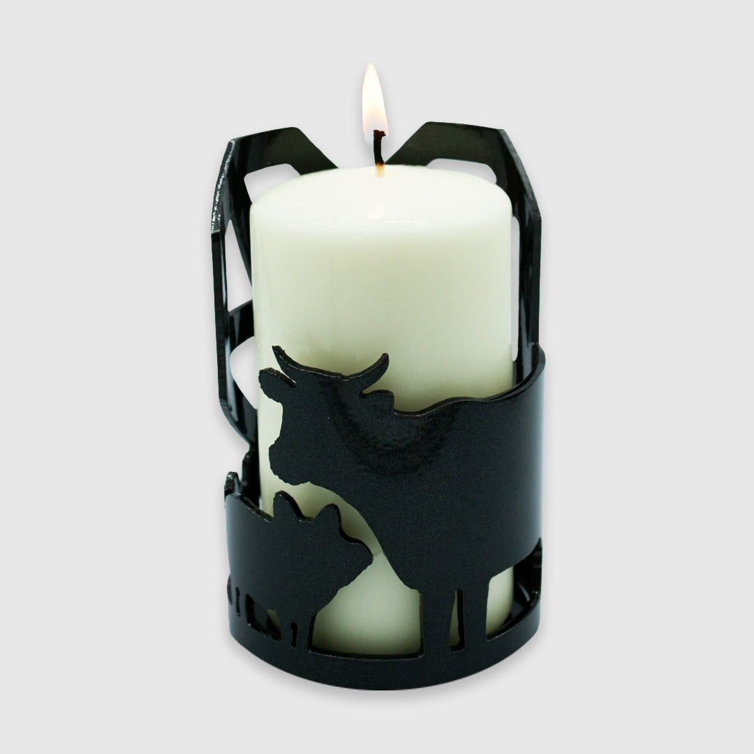 A large white candle that is lit in the candle holder showing the side with the cow and pig cut outs.