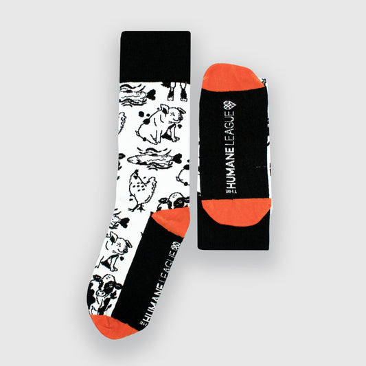 A pair of socks featuring black and white illustrations of cows, pigs, fish, and chickens. The toe and heel are bright red. The top and bottom are solid black. The Humane League logo is on the bottom of the socks.