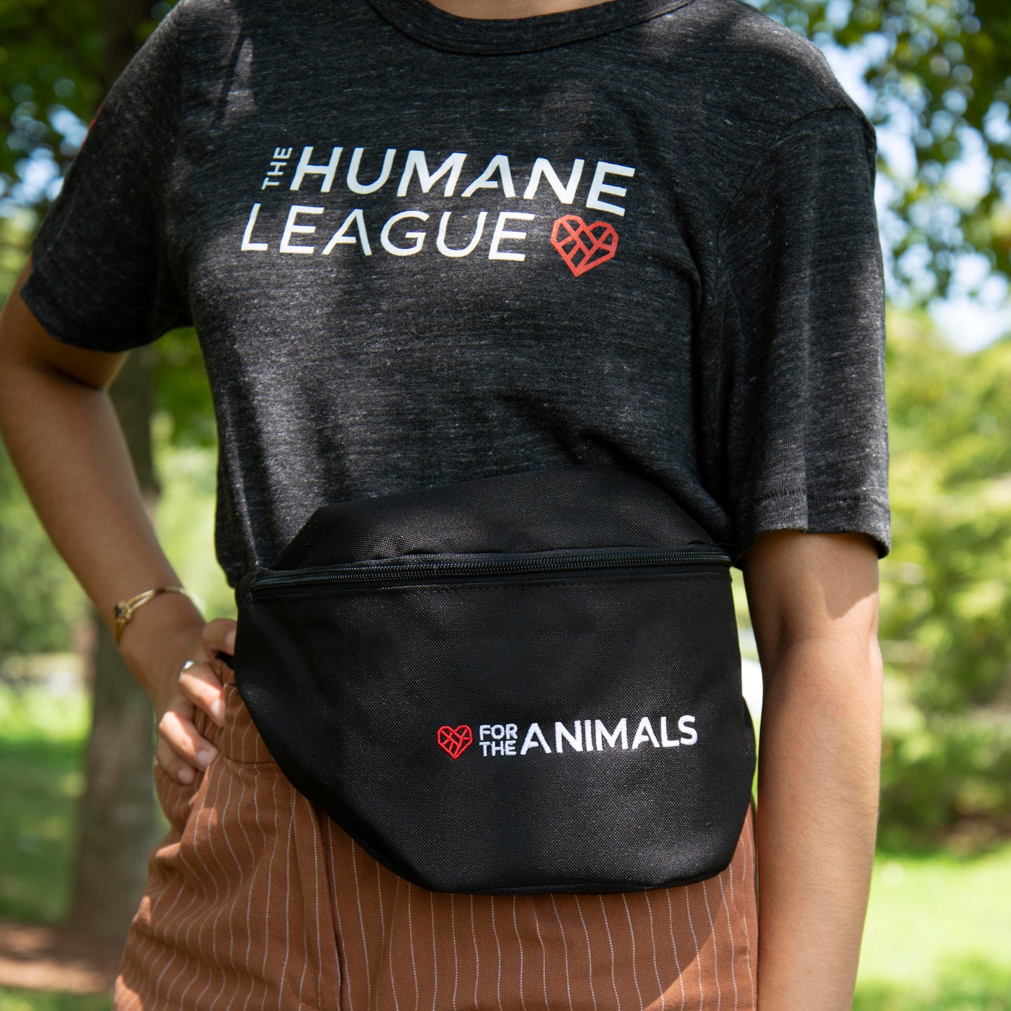 A person wearing a dark gray t-shirt with "The Humane League" logo printed in white and red across the chest, paired with a black fanny pack and brown pinstripe pants. The fanny pack features the slogan "For the Animals" in white text with a small red geometric heart icon. The person is outdoors, with trees and greenery visible in the background.