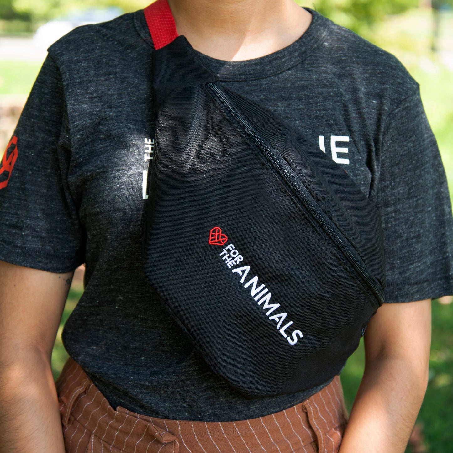 A person wearing a dark gray t-shirt wearing the black fanny pack diagonally across their chest. The fanny pack features the slogan "For the Animals" in white text with a small red geometric heart icon. The person is outdoors, with greenery visible in the background.