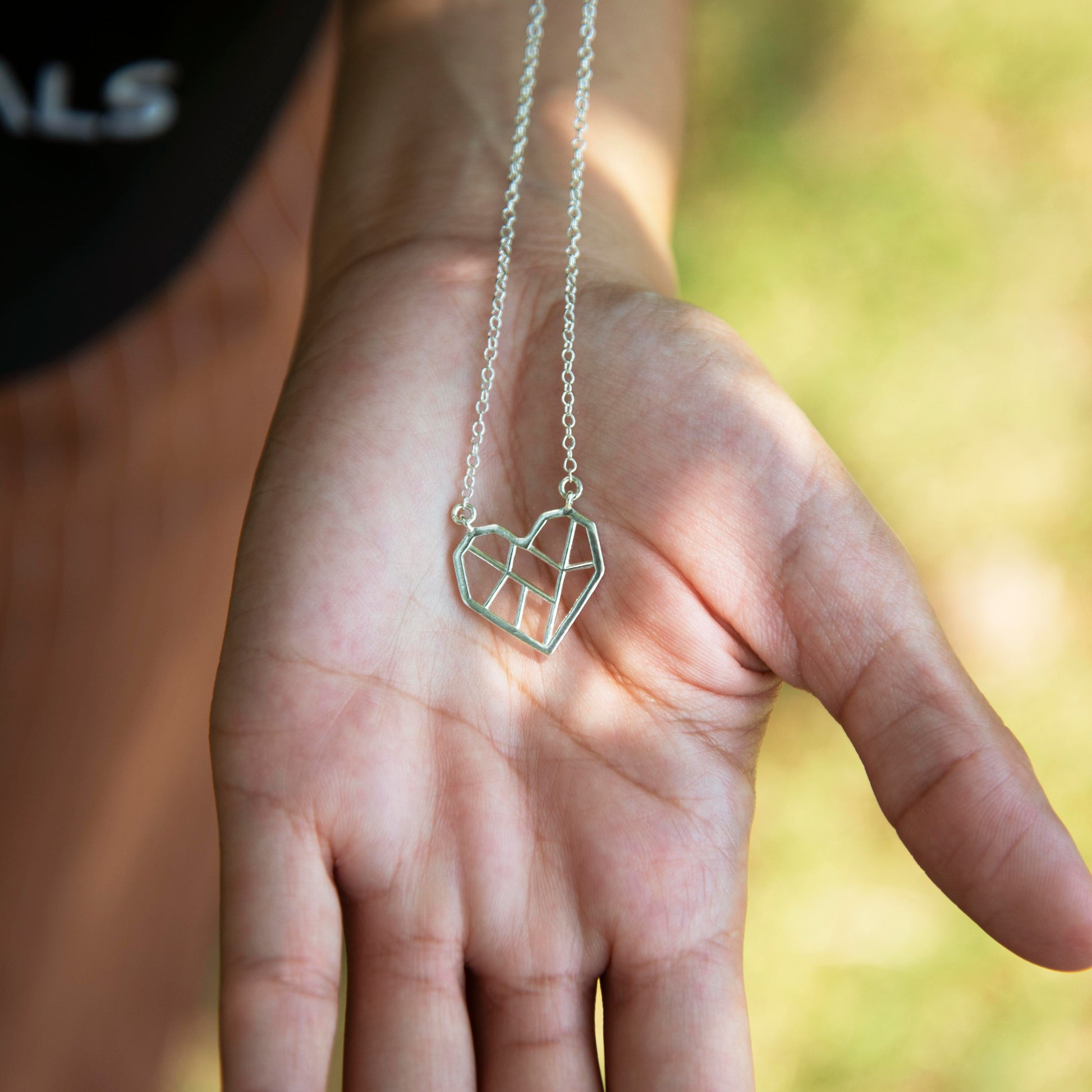 Close up photograph of a person holding the silver heart-shaped pendant in the palm of their hand. The heart pendant is a geometric design featuring clean, angular lines that divide the heart into multiple sections. The pendant hangs from a silver, textured chain that is being pulled away from the pendant and continues out of view. The background is a blurred green outdoor scene.