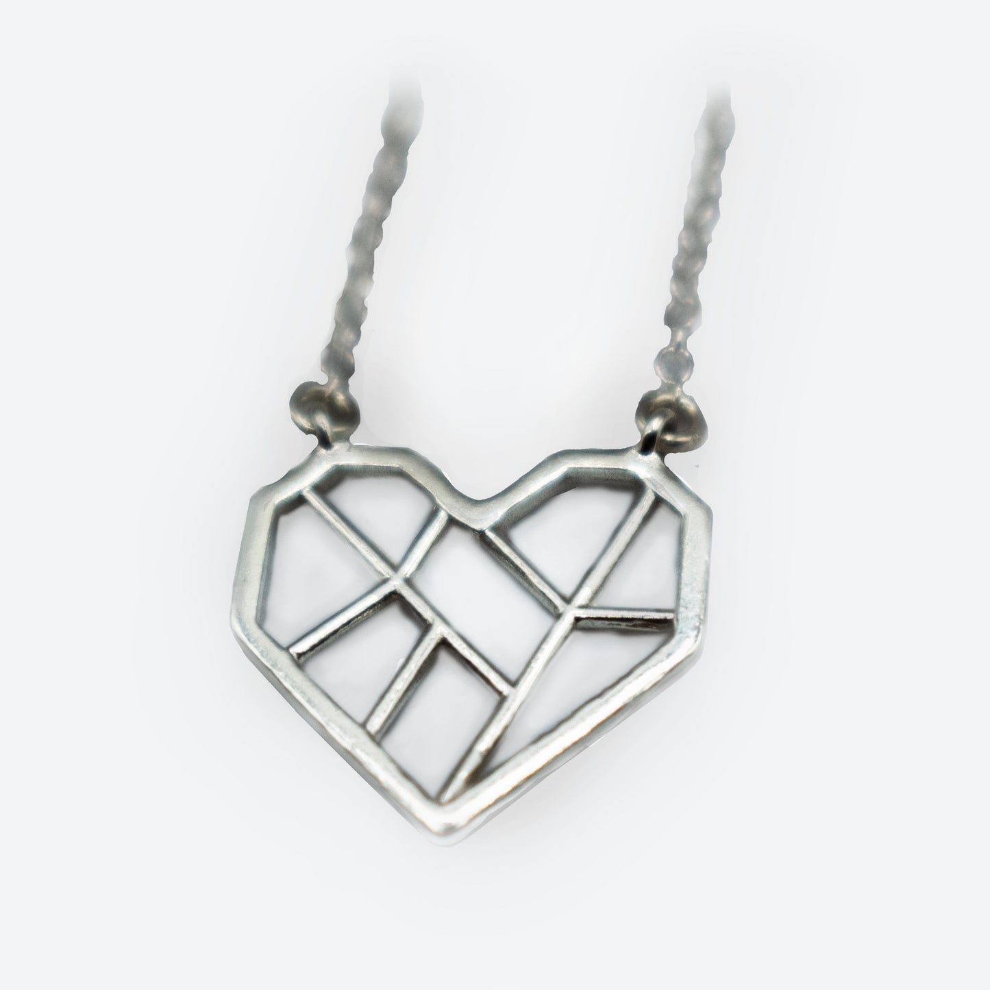 A silver pendant in the shape of a heart, with a geometric design featuring clean, angular lines that divide the heart into multiple sections. The pendant hangs from a silver, textured chain. The overall style is minimalist and modern.
