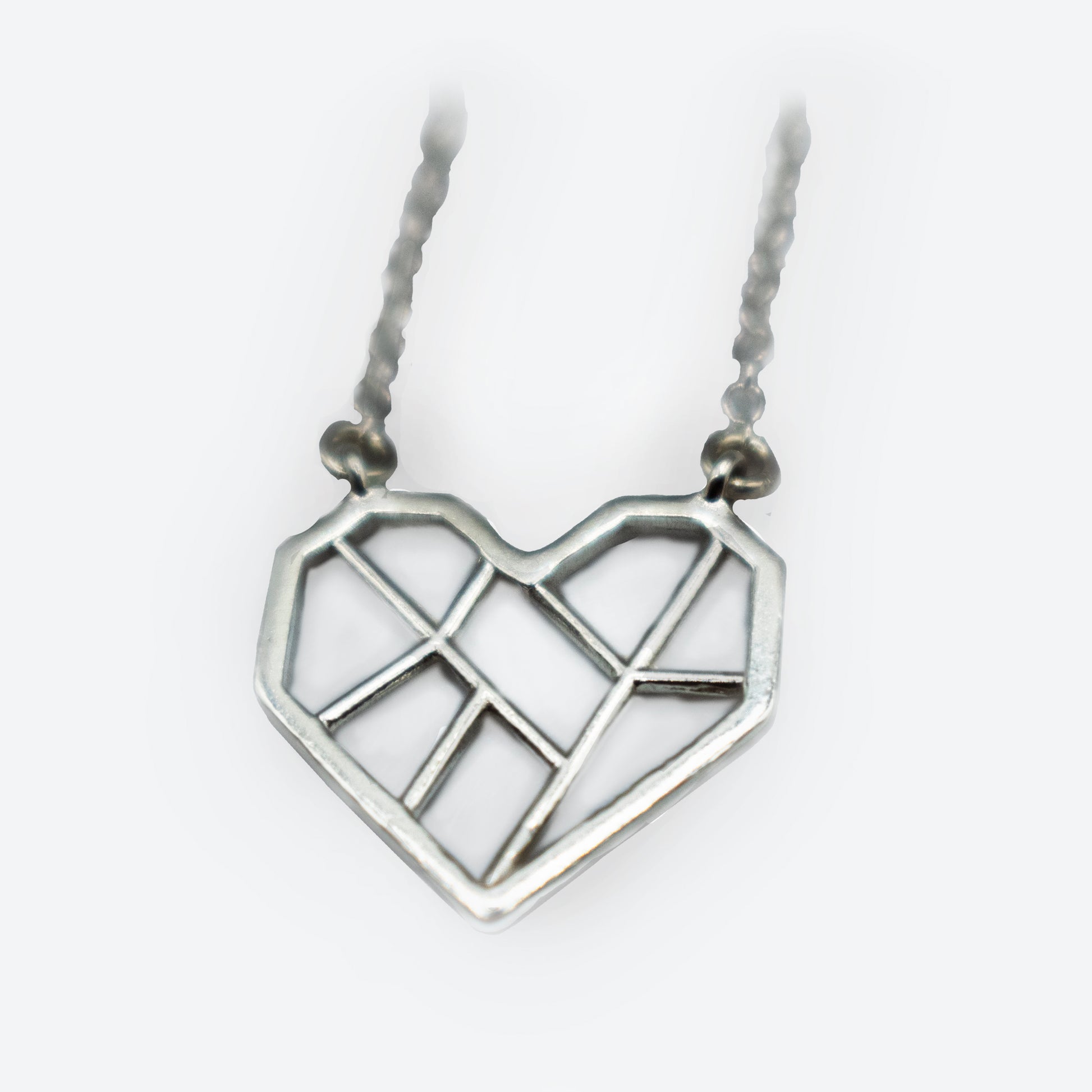 A silver pendant in the shape of a heart, with a geometric design featuring clean, angular lines that divide the heart into multiple sections. The pendant hangs from a silver, textured chain. The overall style is minimalist and modern.